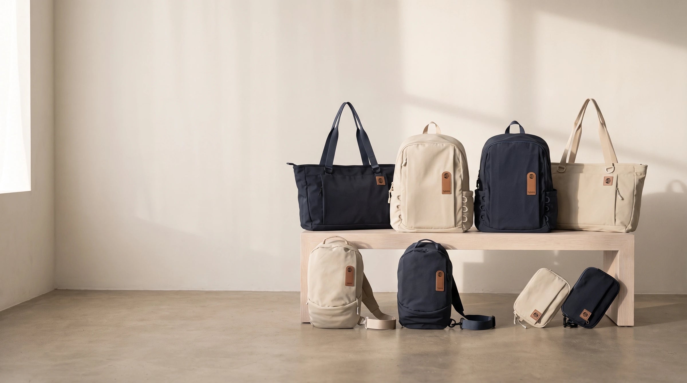 The hybeks Drift collection in Morning Fog and Midnight Transit. Backpack, tote, sling and kit on a pale wood bench.